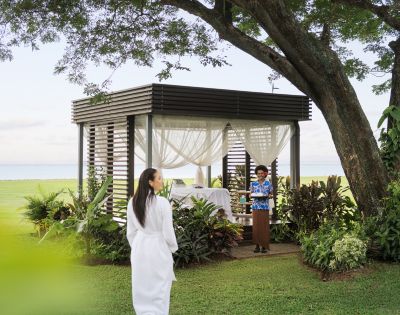 A woman in a white robe walks toward a gazebo with sheer curtains, set in a lush garden by the sea. A staff member in tropical attire awaits with a warm smile.