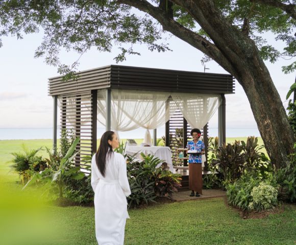 A woman in a white robe walks toward a gazebo with sheer curtains, set in a lush garden by the sea. A staff member in tropical attire awaits with a warm smile.