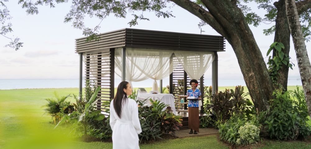 A woman in a white robe walks toward a gazebo with sheer curtains, set in a lush garden by the sea. A staff member in tropical attire awaits with a warm smile.