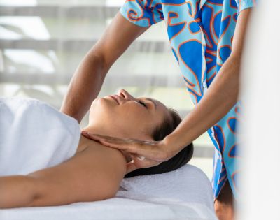 A person receives a relaxing massage, lying with eyes closed and a slight smile. The masseuse, wearing a colorful outfit, gently massages the neck. The atmosphere is calm and serene.