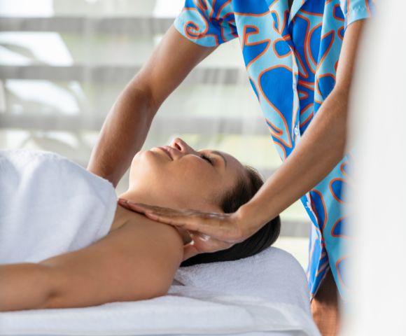 A person receives a relaxing massage, lying with eyes closed and a slight smile. The masseuse, wearing a colorful outfit, gently massages the neck. The atmosphere is calm and serene.