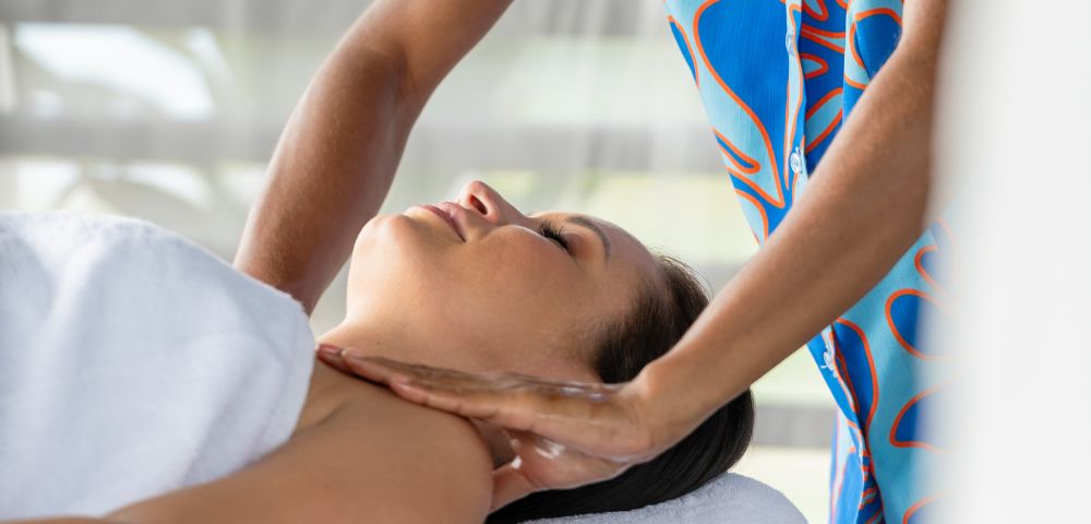 A person receives a relaxing massage, lying with eyes closed and a slight smile. The masseuse, wearing a colorful outfit, gently massages the neck. The atmosphere is calm and serene.