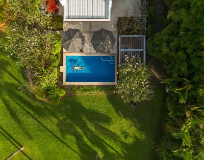 Aerial view of a serene backyard with a woman swimming in a rectangular pool, surrounded by lush greenery and trees casting shadows on a sunny day.