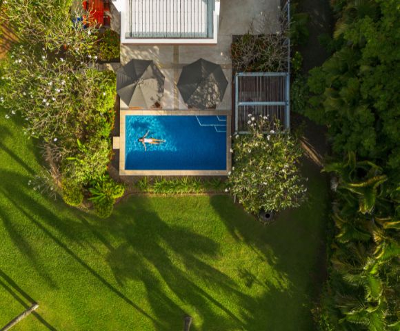 Aerial view of a serene backyard with a woman swimming in a rectangular pool, surrounded by lush greenery and trees casting shadows on a sunny day.
