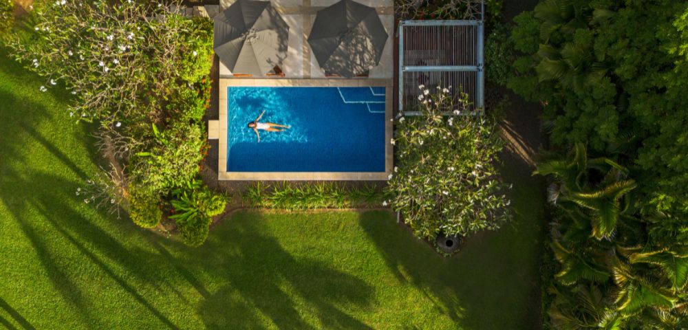 Aerial view of a serene backyard with a woman swimming in a rectangular pool, surrounded by lush greenery and trees casting shadows on a sunny day.