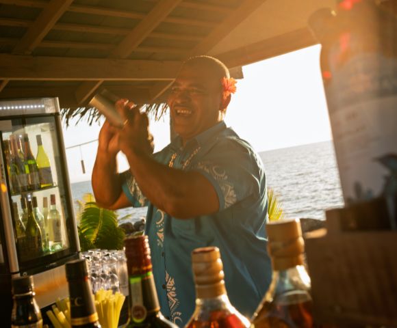 Bartender preparing a cocktail at an outdoor bar with tropical decor, bottles of liquor on the counter, and an ocean view in the background at sunset.