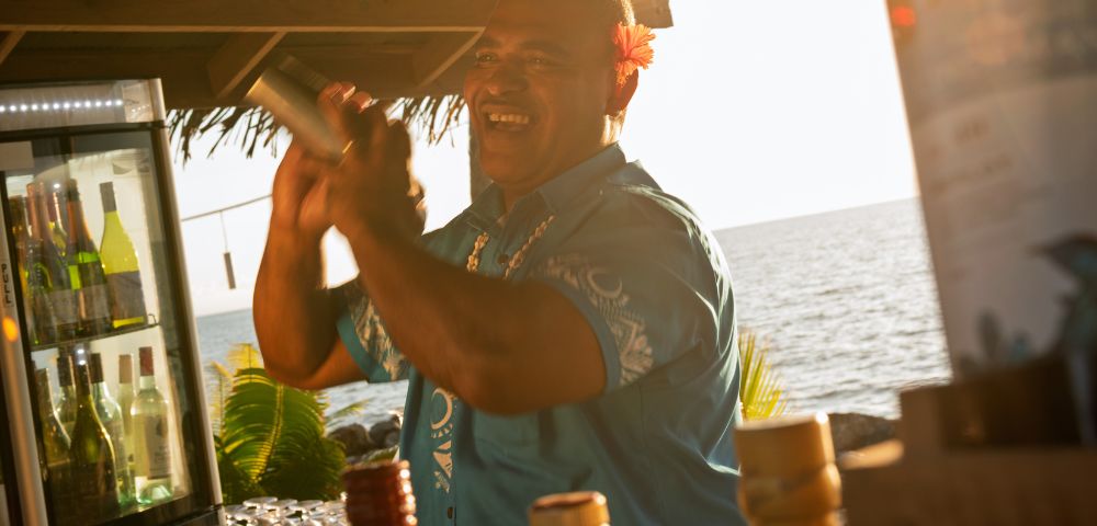 Bartender preparing a cocktail at an outdoor bar with tropical decor, bottles of liquor on the counter, and an ocean view in the background at sunset.