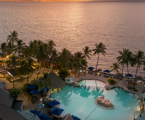 Aerial view of a resort pool surrounded by palm trees at sunset, with the ocean in the background at DoubleTree Resort by Hilton Hotel Fiji.