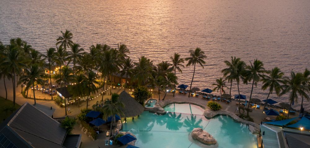 Aerial view of a resort pool surrounded by palm trees at sunset, with the ocean in the background at DoubleTree Resort by Hilton Hotel Fiji.