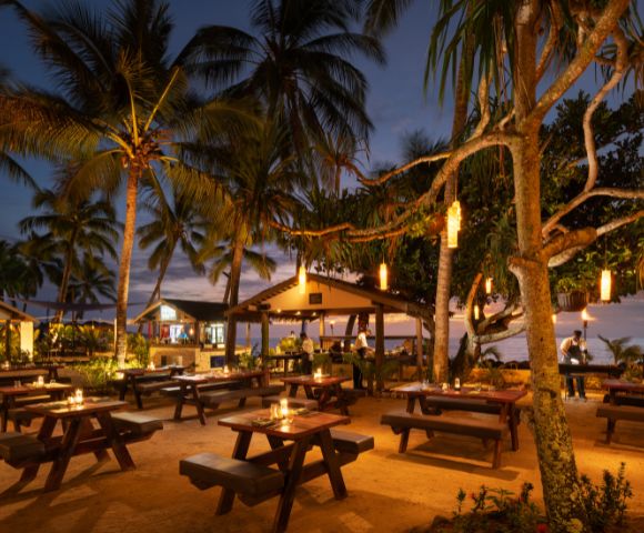 Beachfront dining area illuminated with warm lights and decorated with palm trees, creating a cozy evening ambiance at DoubleTree Resort by Hilton Hotel Fiji.
