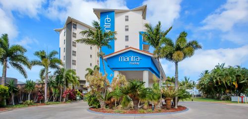 Hotel entrance featuring a blue 'mantra mackay' sign, surrounded by lush tropical plants and palm trees under a bright blue sky.