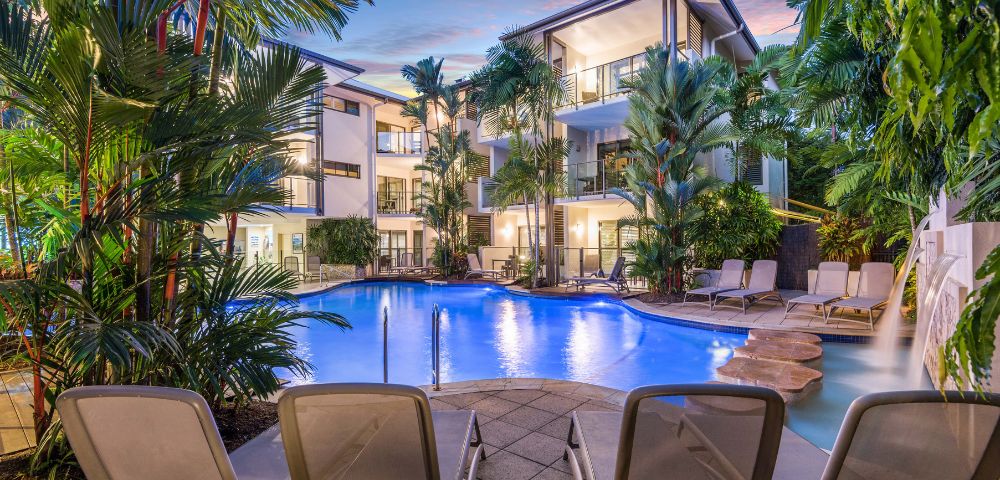Resort pool at night with illuminated water and reflections of palm trees.
