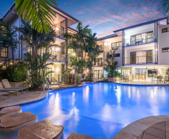 Resort pool at night with illuminated water and reflections of palm trees.