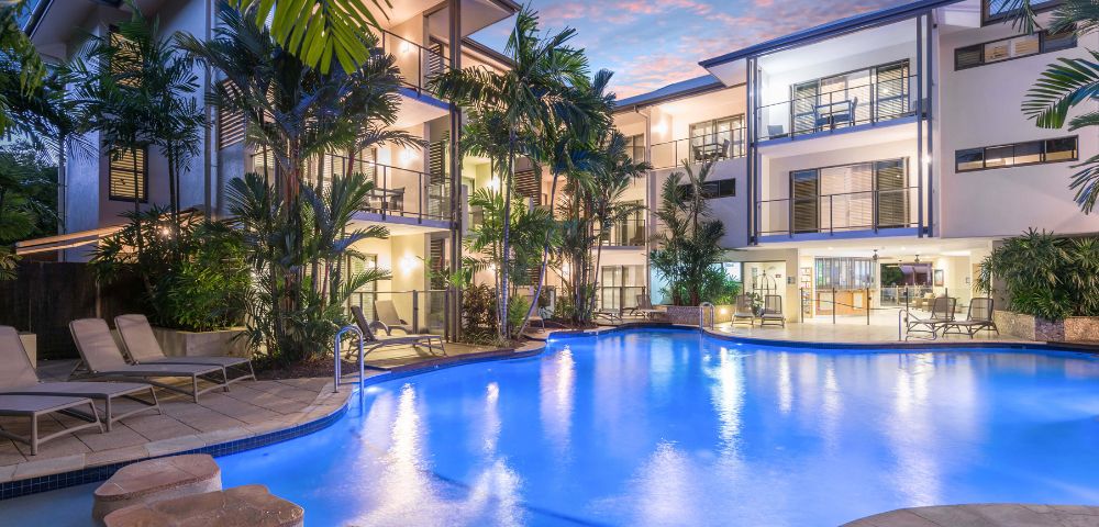 Resort pool at night with illuminated water and reflections of palm trees.