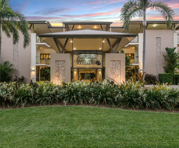 Elegant resort entrance framed by palm trees at sunset, with lush greenery and a welcoming, luxurious ambiance. Sky features vibrant pink and purple hues.