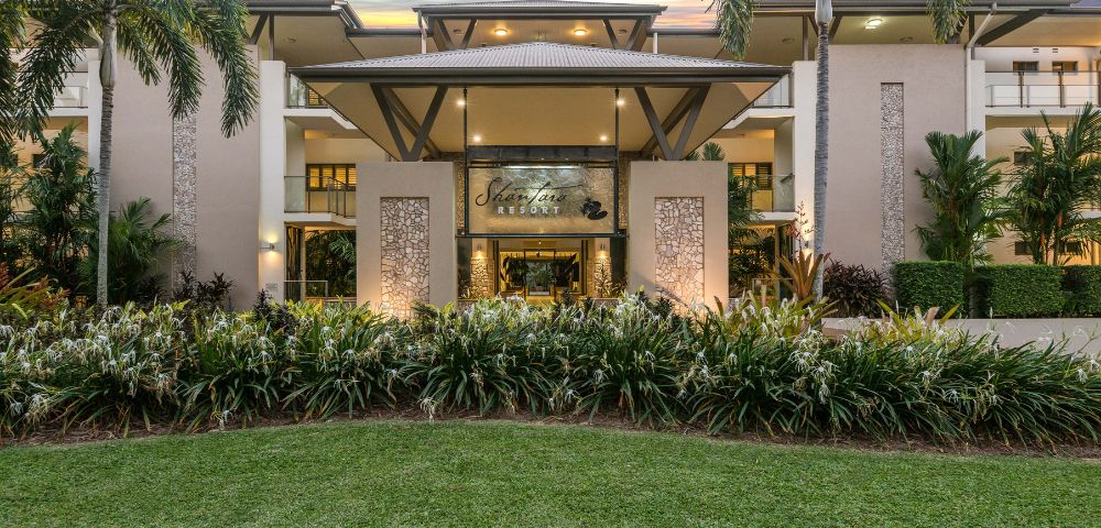 Elegant resort entrance framed by palm trees at sunset, with lush greenery and a welcoming, luxurious ambiance. Sky features vibrant pink and purple hues.