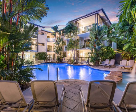 Resort pool at night with illuminated water and reflections of palm trees.