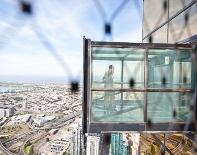 A person stands inside a glass observation box extending from a skyscraper, overlooking a sprawling cityscape. The scene conveys a sense of thrill and awe.