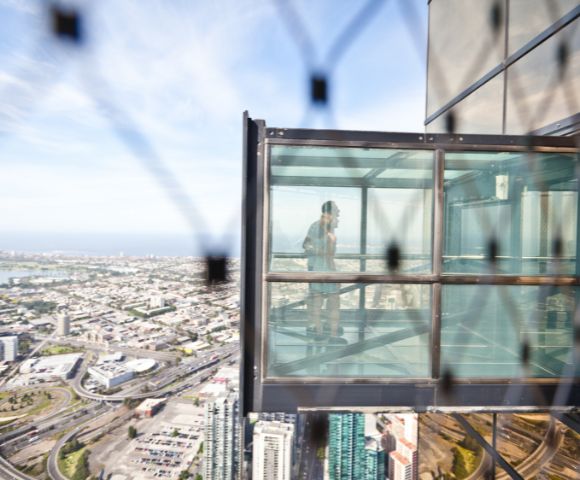 A person stands inside a glass observation box extending from a skyscraper, overlooking a sprawling cityscape. The scene conveys a sense of thrill and awe.