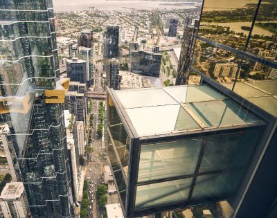 Aerial view of a cityscape with tall glass skyscrapers. A glass observation deck juts out from a building, showing streets below. The atmosphere is modern and urban.