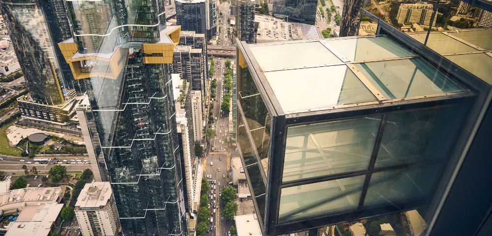 Aerial view of a cityscape with tall glass skyscrapers. A glass observation deck juts out from a building, showing streets below. The atmosphere is modern and urban.