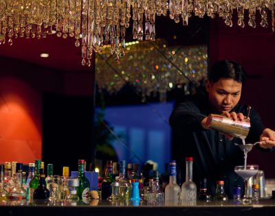 Bartender in a dimly lit bar with red walls, pouring a cocktail into a glass. Bottles line the counter, and a chandelier adds a touch of elegance.