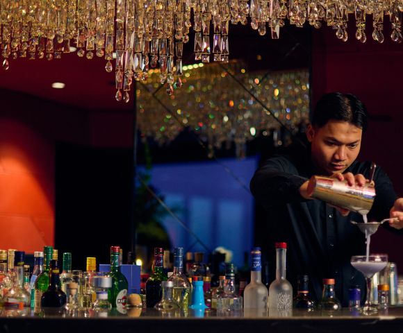 Bartender in a dimly lit bar with red walls, pouring a cocktail into a glass. Bottles line the counter, and a chandelier adds a touch of elegance.