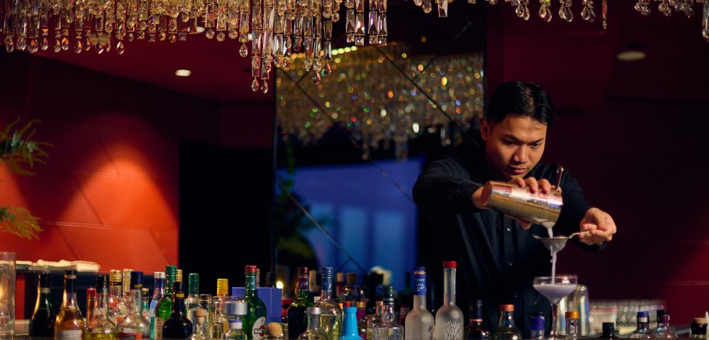 Bartender in a dimly lit bar with red walls, pouring a cocktail into a glass. Bottles line the counter, and a chandelier adds a touch of elegance.