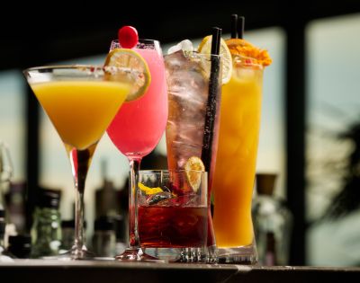 A vibrant array of cocktails, including a yellow martini, pink cocktail, and orange drink, with garnishes, sitting on a bar in a dimly lit setting.