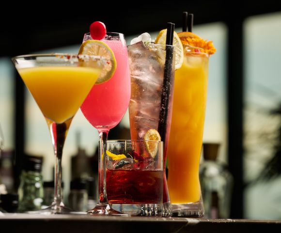 A vibrant array of cocktails, including a yellow martini, pink cocktail, and orange drink, with garnishes, sitting on a bar in a dimly lit setting.