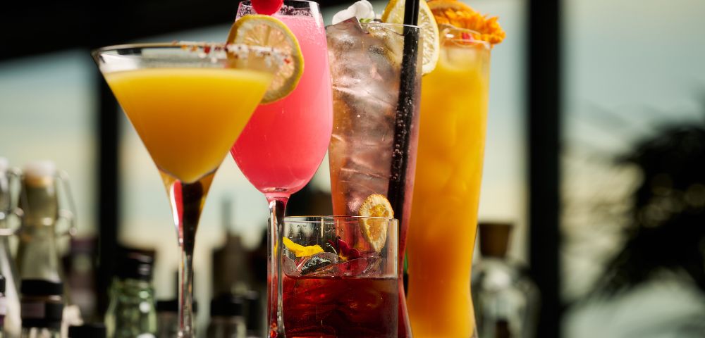 A vibrant array of cocktails, including a yellow martini, pink cocktail, and orange drink, with garnishes, sitting on a bar in a dimly lit setting.