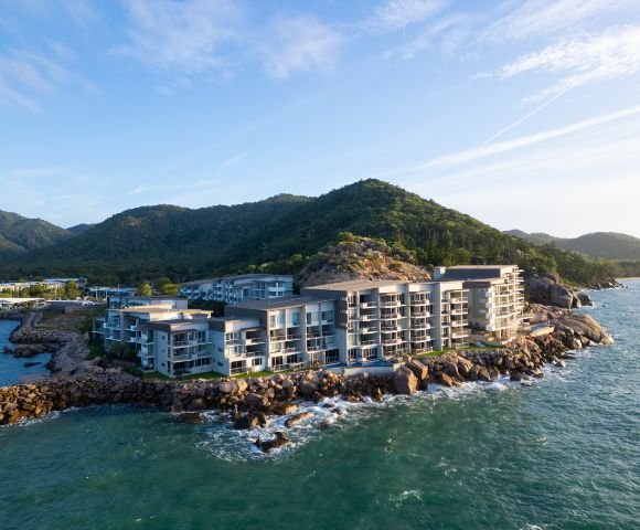 Modern seaside resort on rocky coastline, surrounded by lush green hills and calm ocean waves. Bright, clear day with a serene atmosphere.