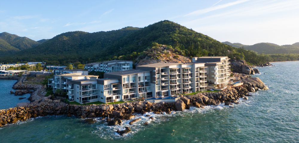 Modern seaside resort on rocky coastline, surrounded by lush green hills and calm ocean waves. Bright, clear day with a serene atmosphere.
