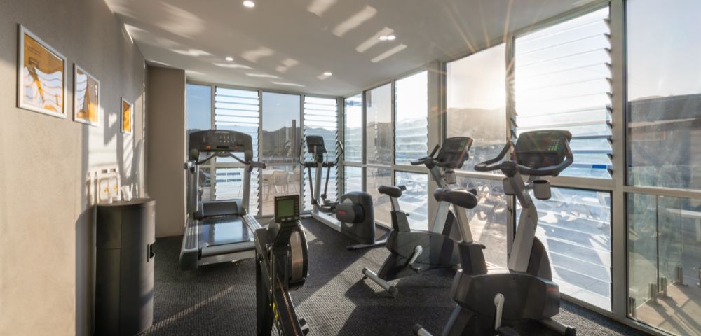 Sunlit gym room with large windows features treadmills, exercise bikes, and a rowing machine. Bright, airy atmosphere with motivational wall art.