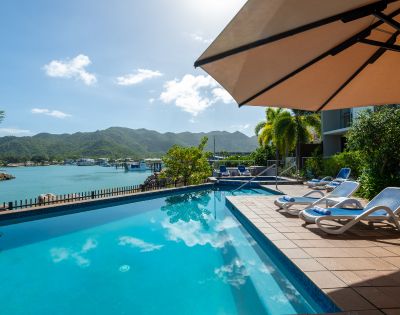 Rooftop pool overlooking a bay with mountains. Lounge chairs line the deck under a large umbrella. Bright, sunny sky reflects in the water. Tranquil ambiance.