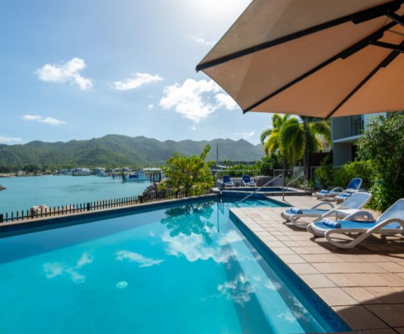 Rooftop pool overlooking a bay with mountains. Lounge chairs line the deck under a large umbrella. Bright, sunny sky reflects in the water. Tranquil ambiance.