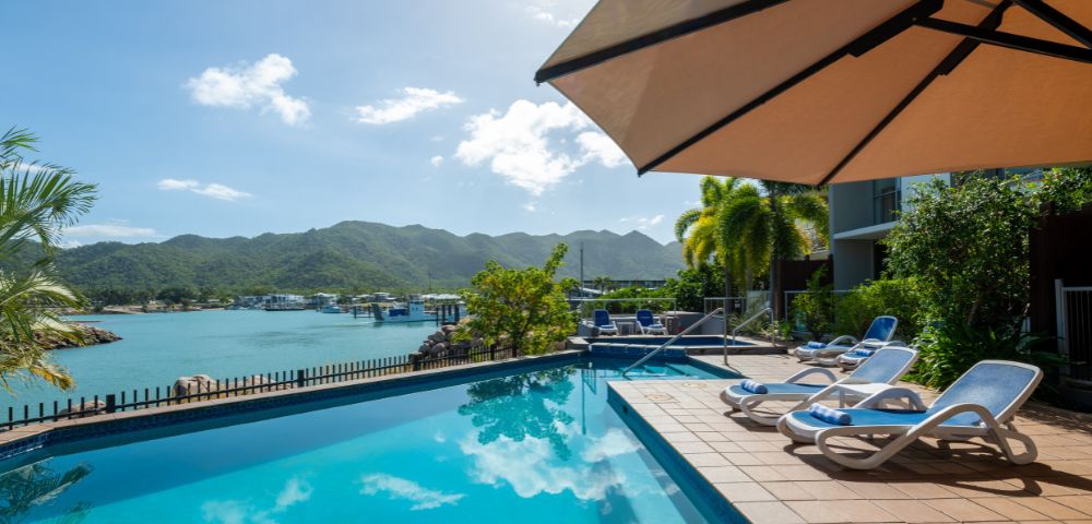 Rooftop pool overlooking a bay with mountains. Lounge chairs line the deck under a large umbrella. Bright, sunny sky reflects in the water. Tranquil ambiance.