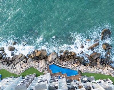 Aerial view of ocean waves crashing on rocky shore beside modern buildings. A uniquely shaped blue pool lies between the structures and the rocks. Tranquil vibe.