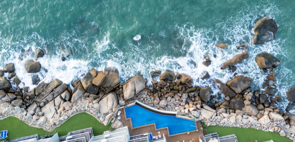 Aerial view of ocean waves crashing on rocky shore beside modern buildings. A uniquely shaped blue pool lies between the structures and the rocks. Tranquil vibe.