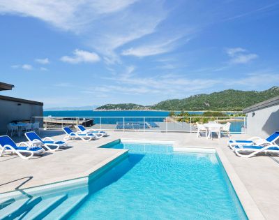 Rooftop terrace with a clear blue pool, surrounded by sun loungers. Overlooks a scenic view of the ocean and green hills under a bright sky.