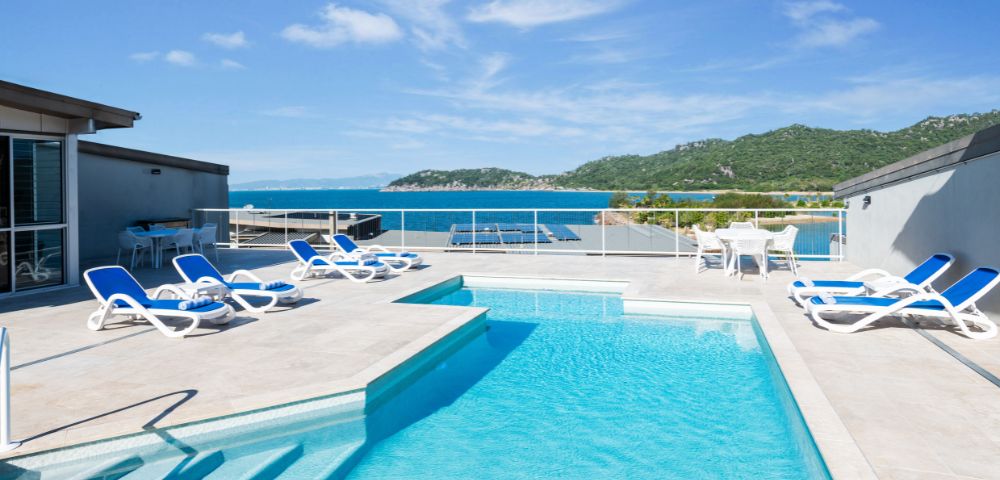 Rooftop terrace with a clear blue pool, surrounded by sun loungers. Overlooks a scenic view of the ocean and green hills under a bright sky.
