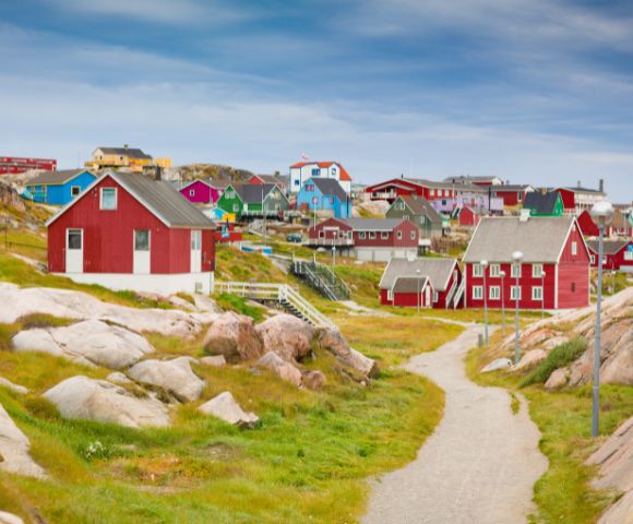A scenic view of a Greenland village features colorful wooden houses, mainly red, scattered across rocky, grassy terrain under a cloudy sky.