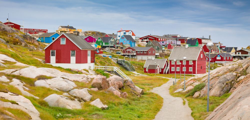 A scenic view of a Greenland village features colorful wooden houses, mainly red, scattered across rocky, grassy terrain under a cloudy sky.