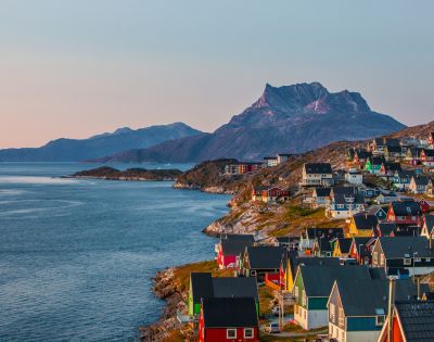 Coastal town at sunset with colorful houses on a hillside, a calm blue sea, and rugged mountains in the background, conveying a serene atmosphere.