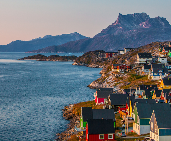 Coastal town at sunset with colorful houses on a hillside, a calm blue sea, and rugged mountains in the background, conveying a serene atmosphere.