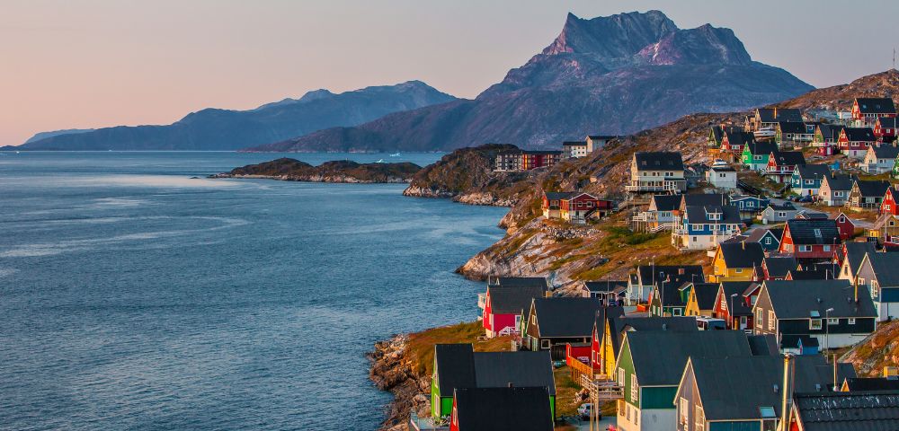 Coastal town at sunset with colorful houses on a hillside, a calm blue sea, and rugged mountains in the background, conveying a serene atmosphere.