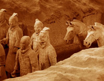 Ancient terracotta warrior statues stand in rows inside an excavation site. The earthy tones of the figures and surrounding soil highlight their historical significance.