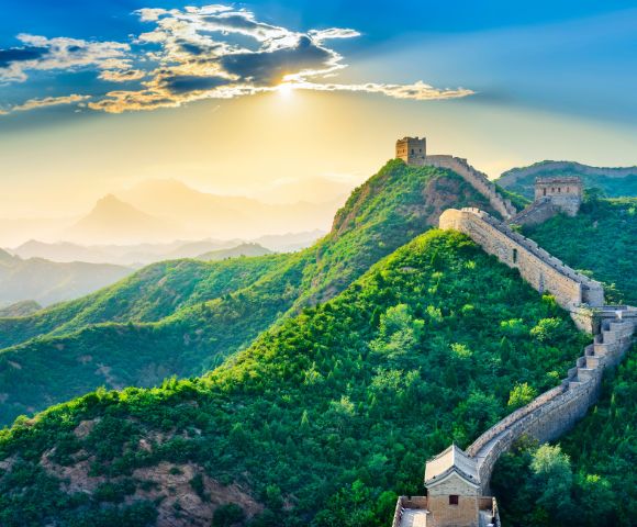 The Great Wall of China winds across lush green mountains under a bright sky. Its stone structure stretches into the distance, showcasing the scale and grandeur of this historic landmark.