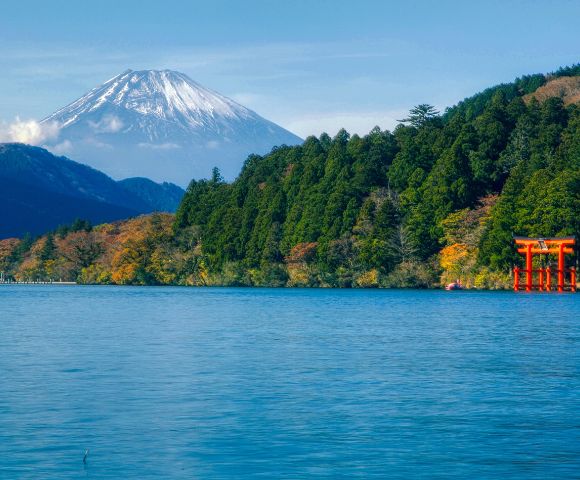 A serene blue lake stretches toward a forested shoreline with a red torii gate standing near the water’s edge. Snow-capped mountains rise in the background under a clear blue sky.