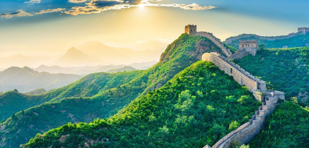 The Great Wall of China winds across lush green mountains under a bright sky. Its stone structure stretches into the distance, showcasing the scale and grandeur of this historic landmark.